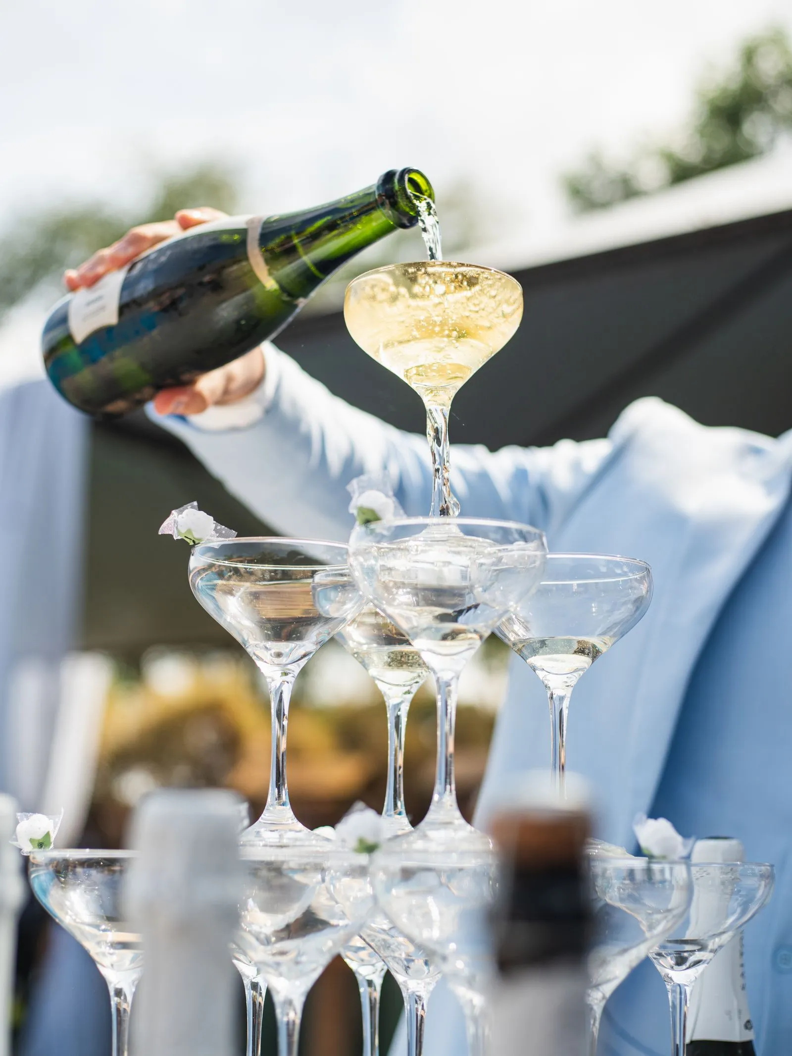 Sparkling wine being poured into a tiered tower of coupe glasses at an outdoor celebration