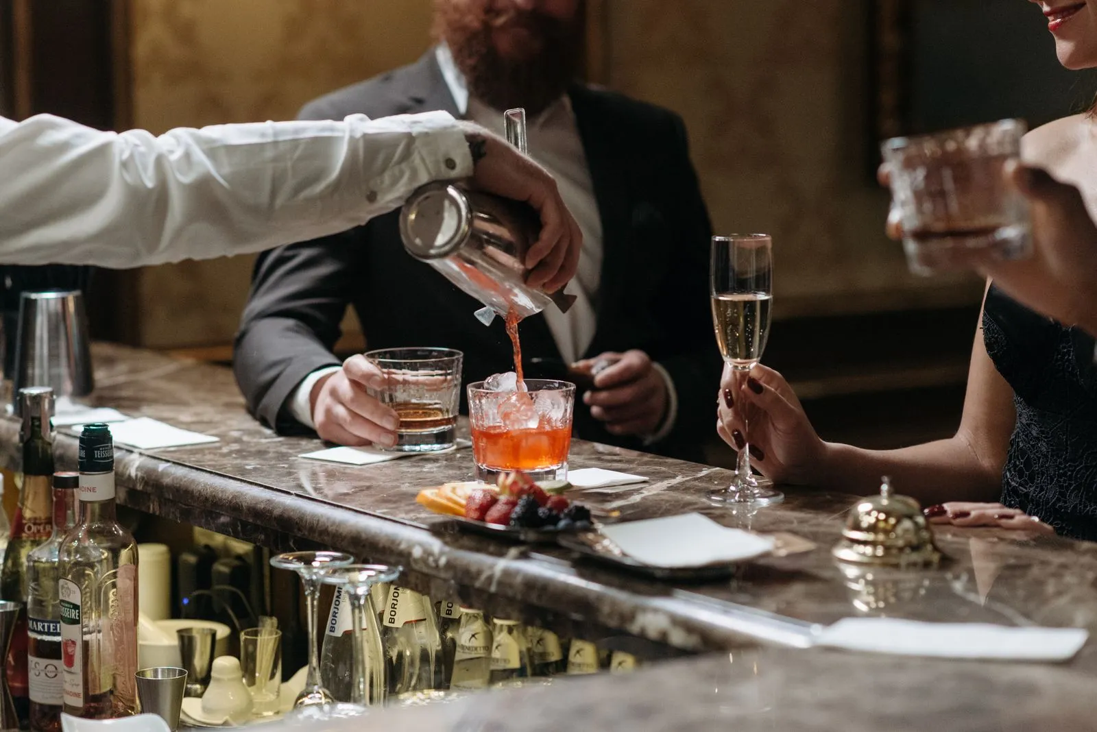 A bartender pouring a cocktail for well-dressed guests along a marble bar counter