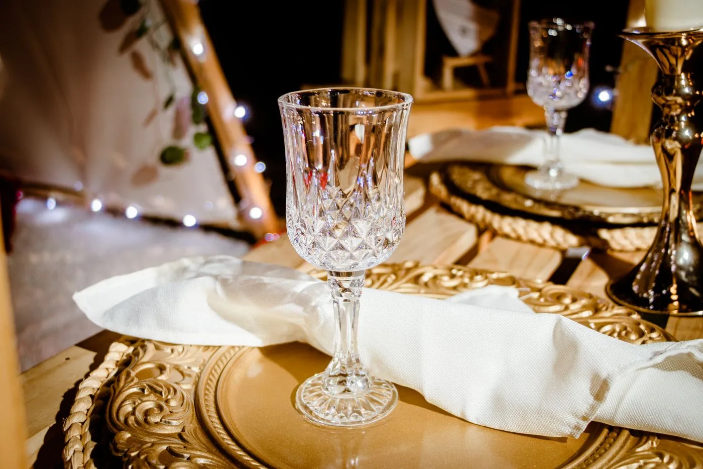 A place setting with a crystal glass on a gold charger plate beside a lit candle