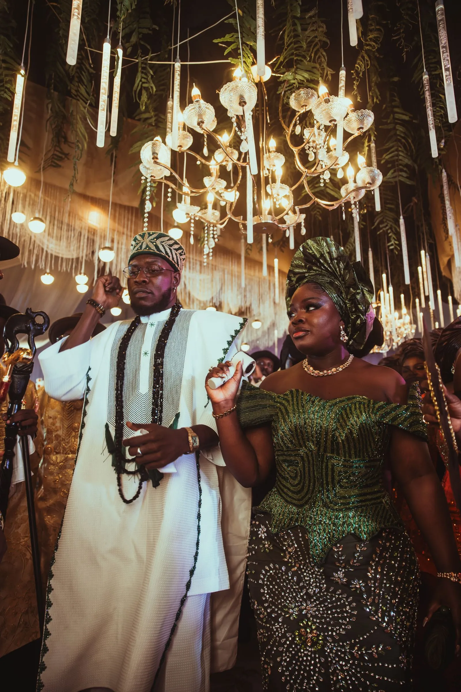 A couple in traditional ceremonial dress in front of a gold-lit celebration backdrop