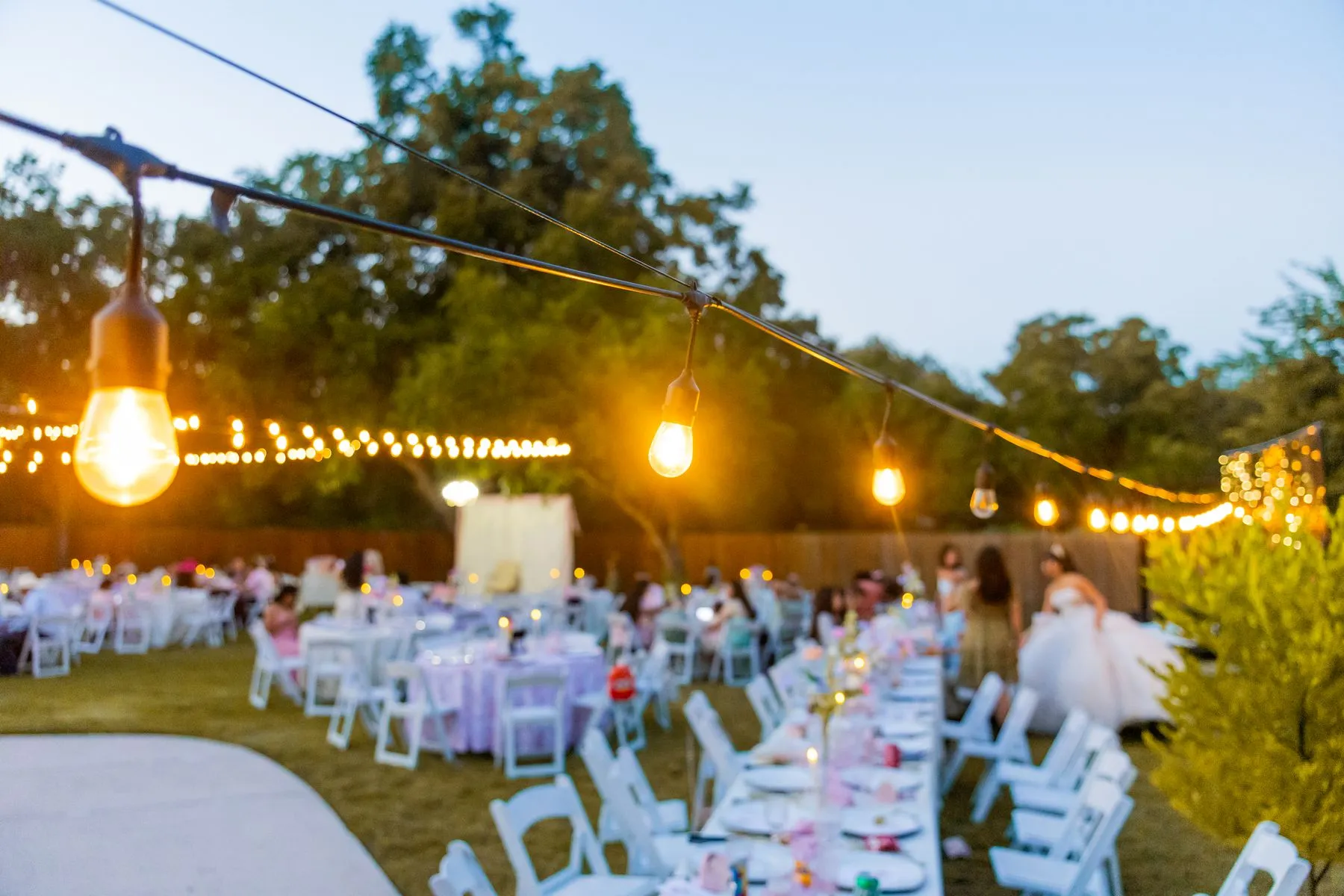 An outdoor wedding reception at dusk with long dressed tables under strung lights