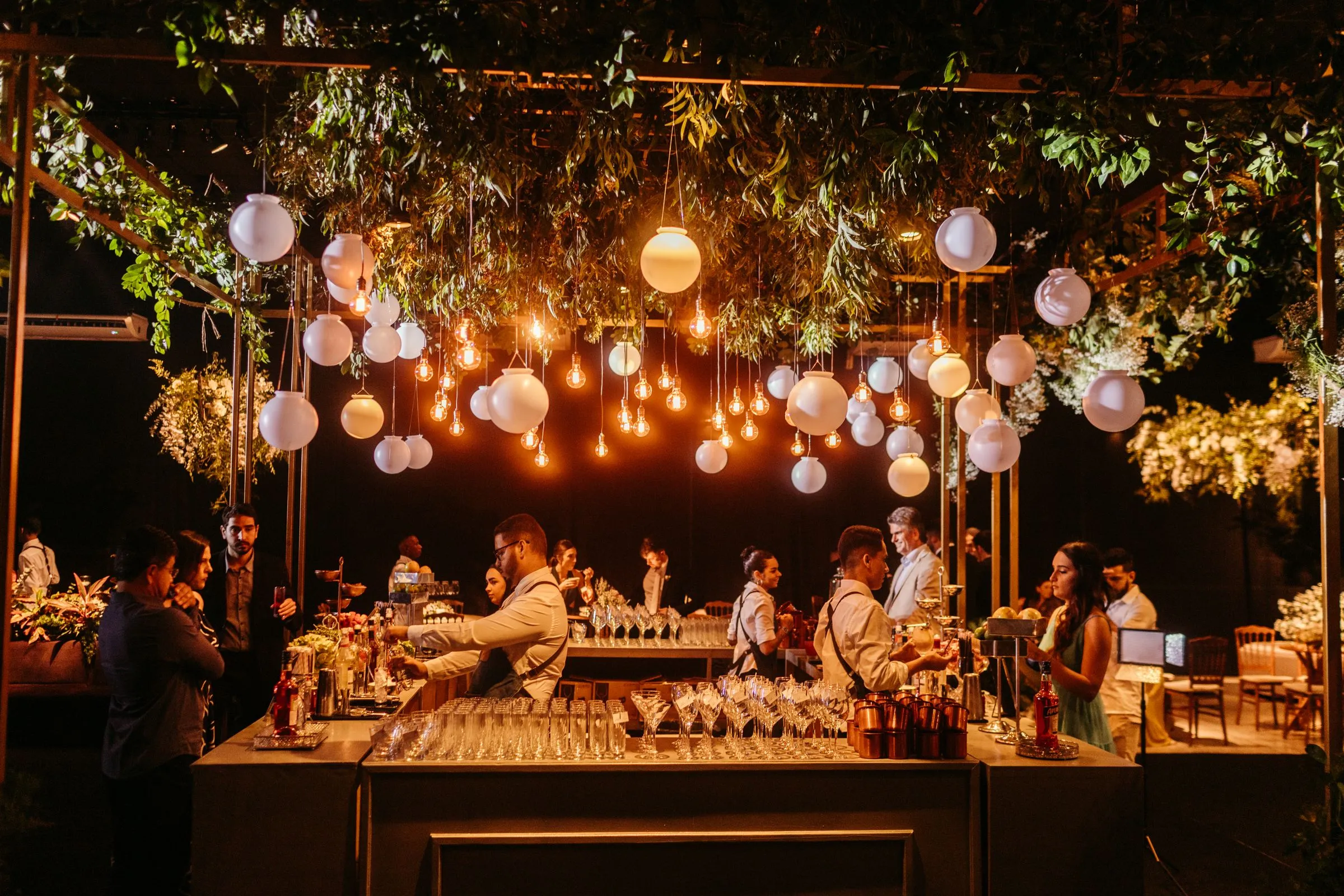 A styled event bar hung with pendant lights and greenery, guests gathered in front of it at dusk
