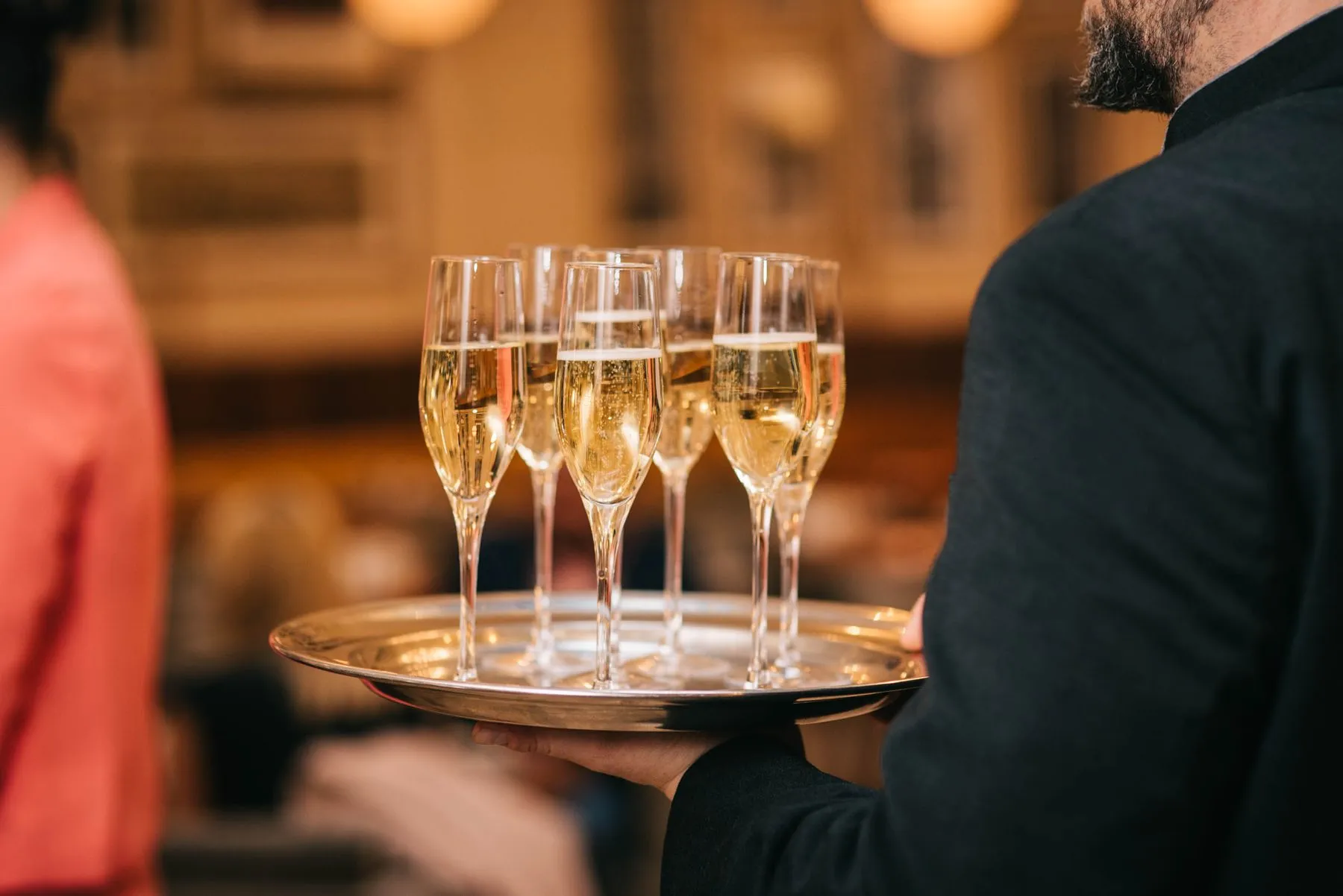 A server carrying a silver tray of filled sparkling wine glasses through an event